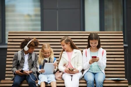 Four children sit on a wooden bench, each engaged with electronic devices. From left to right: a boy with headphones and a mobile, a girl with two plaits using a tablet, a girl with a mobile, and a girl in a white jacket looking at a smartphone.