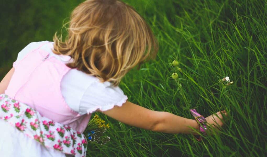 A young child with blonde hair, wearing a white shirt and a floral pink dress, is crouched down and reaching into tall, green grass, appearing to pick flowers or explore the plants on a bright, sunny day.
