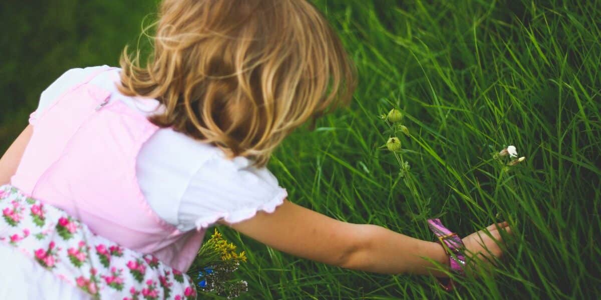 A young child with blonde hair, wearing a white shirt and a floral pink dress, is crouched down and reaching into tall, green grass, appearing to pick flowers or explore the plants on a bright, sunny day.