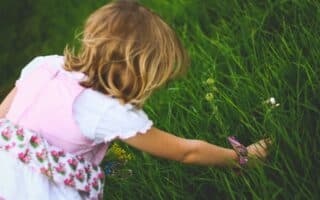 A young child with blonde hair, wearing a white shirt and a floral pink dress, is crouched down and reaching into tall, green grass, appearing to pick flowers or explore the plants on a bright, sunny day.