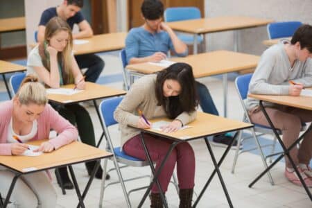 A group of students sits at individual desks in a classroom, focused on writing or taking an exam. The students appear concentrated, each with a pen and paper, and the classroom has a bright, orderly atmosphere.