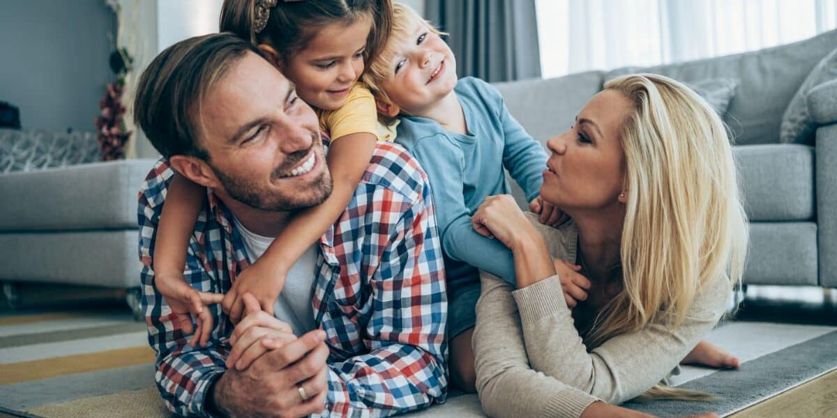A smiling family of four lies playfully on the living room floor. The father and mother face each other, smiling, whilst their two young children, a girl and a boy, climb on their backs and laugh. The room is bright with large windows and cosy decor.