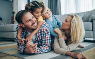 A smiling family of four lies playfully on the living room floor. The father and mother face each other, smiling, whilst their two young children, a girl and a boy, climb on their backs and laugh. The room is bright with large windows and cosy decor.
