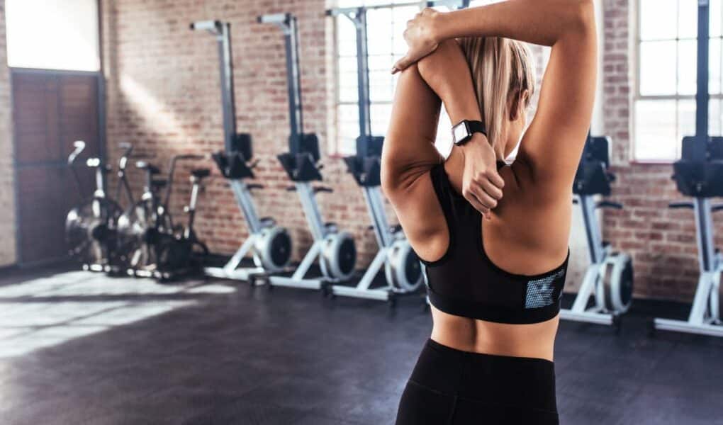 A woman wearing a black sports bra and leggings stretches her arm across her chest in a gym. She faces away from the camera, and exercise bikes and rowing machines are lined up against brick walls with large windows in the background.