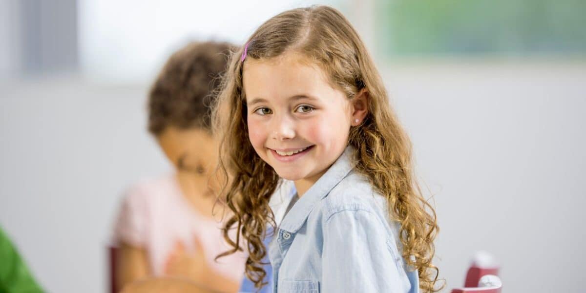 A young girl with long curly hair and a light blue shirt sits at a table, smiling at the camera. Another child, slightly out of focus, sits beside her, focused on colourful clay. The background is bright and blurred.