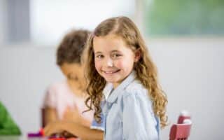 A young girl with long curly hair and a light blue shirt sits at a table, smiling at the camera. Another child, slightly out of focus, sits beside her, focused on colourful clay. The background is bright and blurred.