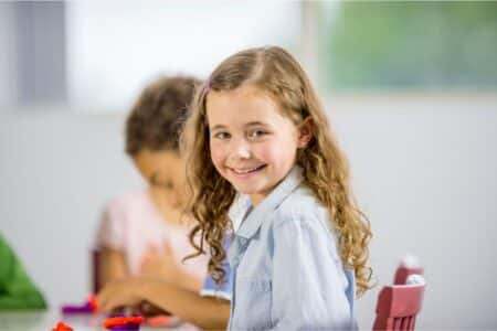 A young girl with long curly hair and a light blue shirt sits at a table, smiling at the camera. Another child, slightly out of focus, sits beside her, focused on colourful clay. The background is bright and blurred.