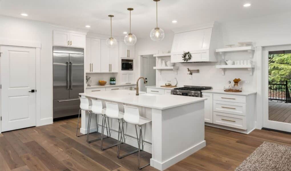 Modern kitchen with white cupboards and island, gold fittings, stainless steel fridge, and gas hob. Four white stools sit at the island, which has a sink and gold tap. Glass pendant lights hang above. Wooden floors and large window showing greenery outside.