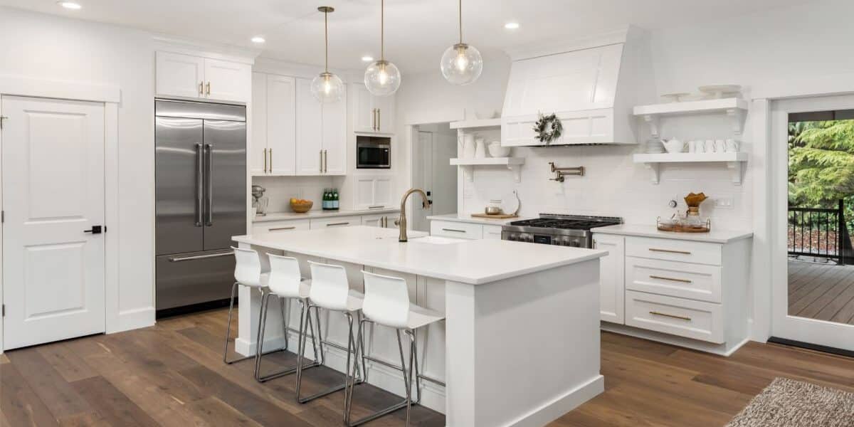 Modern kitchen with white cupboards and island, gold fittings, stainless steel fridge, and gas hob. Four white stools sit at the island, which has a sink and gold tap. Glass pendant lights hang above. Wooden floors and large window showing greenery outside.