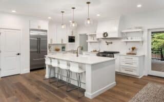 Modern kitchen with white cupboards and island, gold fittings, stainless steel fridge, and gas hob. Four white stools sit at the island, which has a sink and gold tap. Glass pendant lights hang above. Wooden floors and large window showing greenery outside.