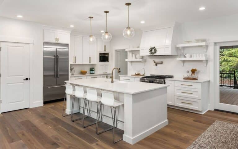 Modern kitchen with white cupboards and island, gold fittings, stainless steel fridge, and gas hob. Four white stools sit at the island, which has a sink and gold tap. Glass pendant lights hang above. Wooden floors and large window showing greenery outside.