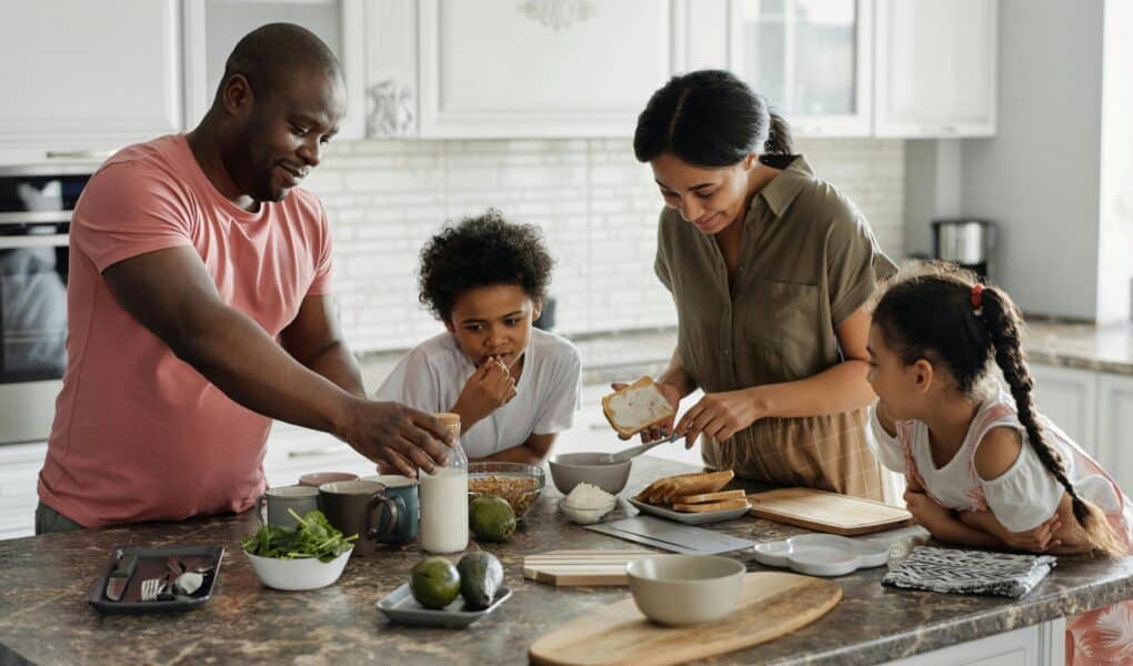 A family of four prepares food together in a bright kitchen. The father reaches for ingredients, the mother spreads something on bread, while their two young children watch and help at the counter with plates, vegetables, and milk. Everyone looks engaged and happy.