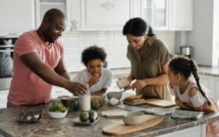 A family of four prepares food together in a bright kitchen. The father reaches for ingredients, the mother spreads something on bread, while their two young children watch and help at the counter with plates, vegetables, and milk. Everyone looks engaged and happy.