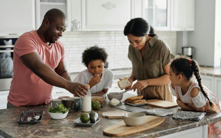 A family of four prepares food together in a bright kitchen. The father reaches for ingredients, the mother spreads something on bread, while their two young children watch and help at the counter with plates, vegetables, and milk. Everyone looks engaged and happy.