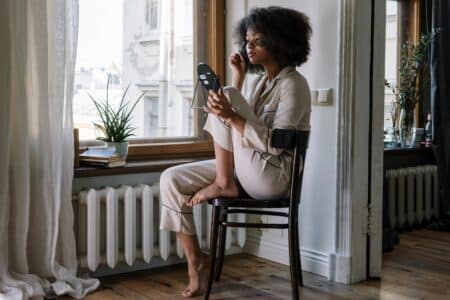A woman in light pyjamas sits cross-legged on a chair by a window, holding a small mirror and applying make-up. She is barefoot, and natural light fills the room. There are books and a potted plant on the windowsill beside her.
