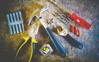 A variety of tools on a worn metal surface: a hammer, pliers, tape measure, adjustable spanners, red screwdriver with bits, wall plugs, screws, and nails, arranged in a semi-circle and lit brightly from above.