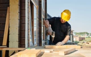 A construction worker wearing a yellow hard hat and gloves drills into a wooden plank at an outdoor building site, with unfinished wooden beams and tools on the table, beside a partially constructed house under a clear sky.
