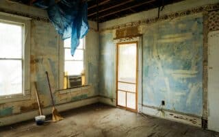 An old, run-down room with peeling blue paint on the walls, exposed ceiling beams, two windows, an air conditioner, and a wooden door. A broom, mop and bucket rest on the worn wooden floor; a blue tarpaulin hangs from the ceiling.