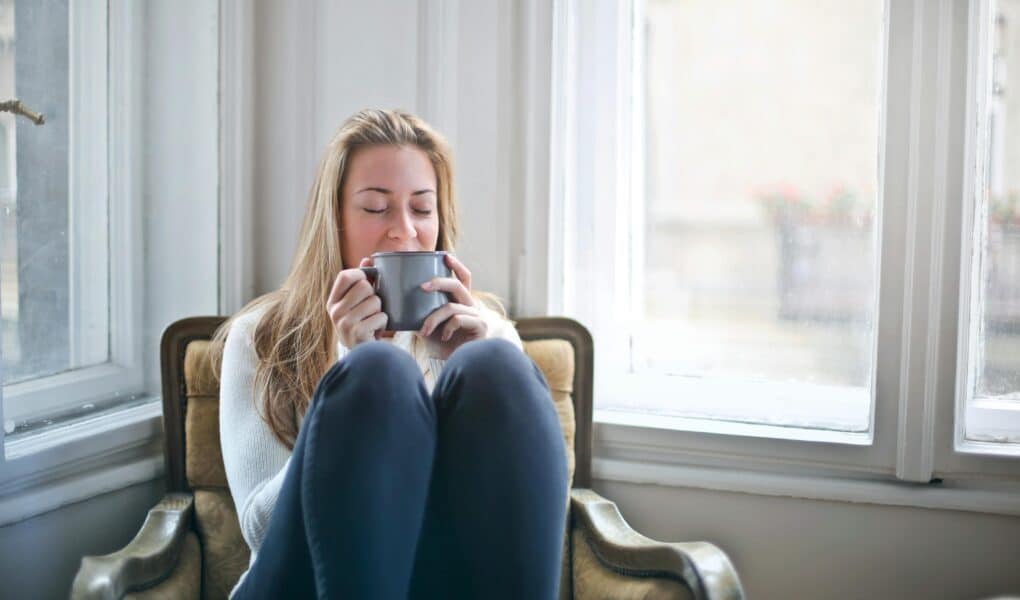 A woman with long blonde hair sits curled up on a cushioned armchair by a bright window, wearing a light jumper and dark trousers, holding a grey mug with both hands near her face, eyes closed, appearing relaxed and content.