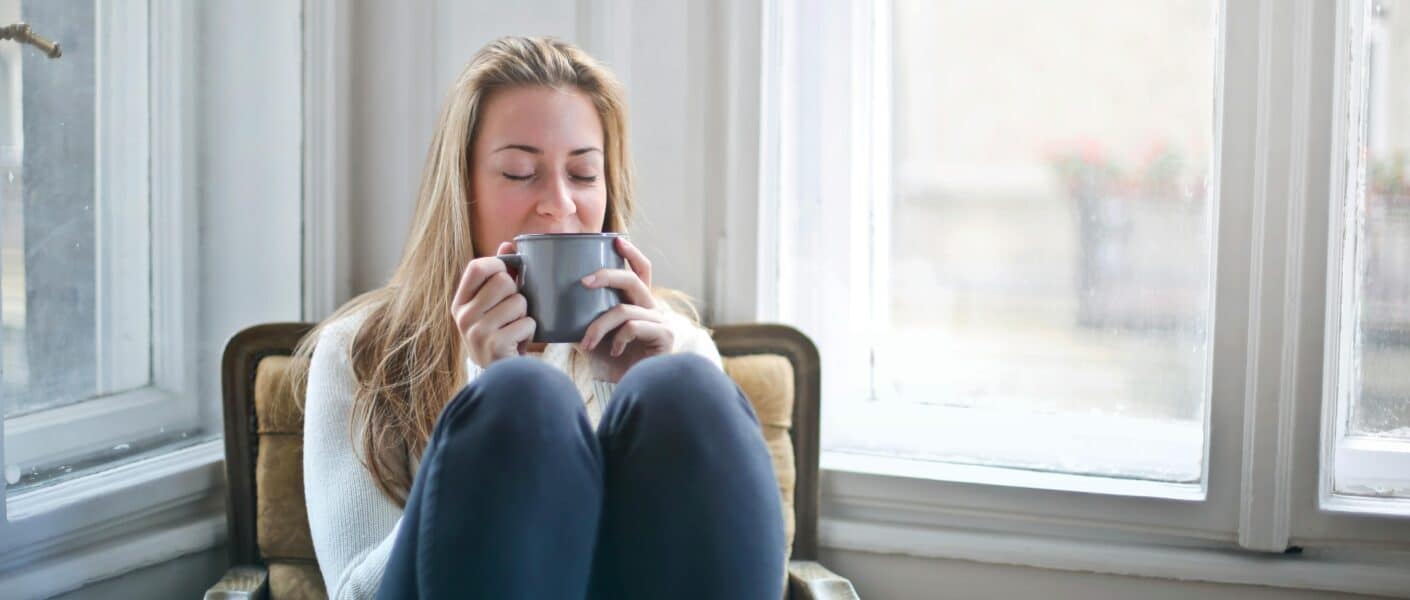 A woman with long blonde hair sits curled up on a cushioned armchair by a bright window, wearing a light jumper and dark trousers, holding a grey mug with both hands near her face, eyes closed, appearing relaxed and content.