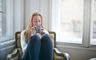 A woman with long blonde hair sits curled up on a cushioned armchair by a bright window, wearing a light jumper and dark trousers, holding a grey mug with both hands near her face, eyes closed, appearing relaxed and content.