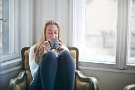 A woman with long blonde hair sits curled up on a cushioned armchair by a bright window, wearing a light jumper and dark trousers, holding a grey mug with both hands near her face, eyes closed, appearing relaxed and content.