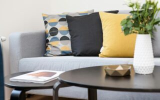 A modern living room features a light grey sofa with three decorative cushions—one geometric, one black, and one yellow. In the foreground, there are two black nesting tables holding a white vase with white flowers, a gold bowl, and a magazine.