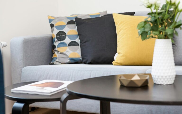 A modern living room features a light grey sofa with three decorative cushions—one geometric, one black, and one yellow. In the foreground, there are two black nesting tables holding a white vase with white flowers, a gold bowl, and a magazine.