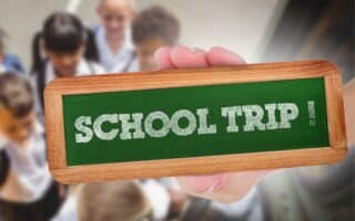 A hand holds a small green chalkboard with the words “SCHOOL TRIP!” written in white chalk. In the blurred background, a group of children in school uniforms gather, suggesting excitement and anticipation for the trip.