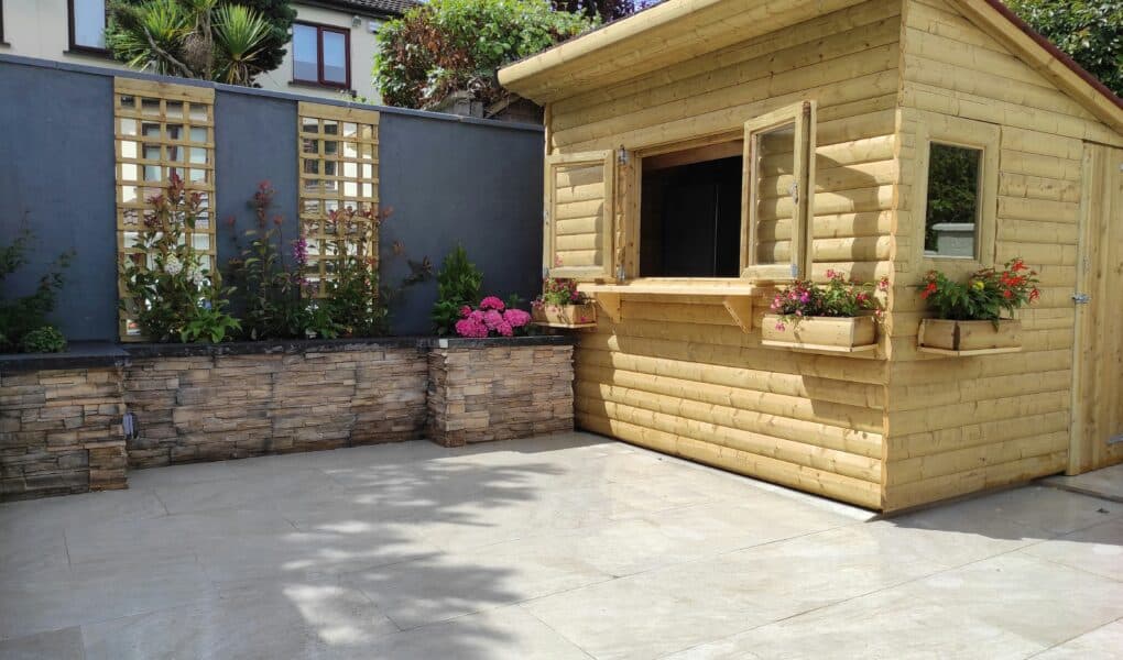 A tidy patio with light stone tiles, a raised garden bed with flowers and shrubs along a grey wall, and a wooden shed with open windows and flower boxes, under a partly sunny sky. A house and trees are visible in the background.