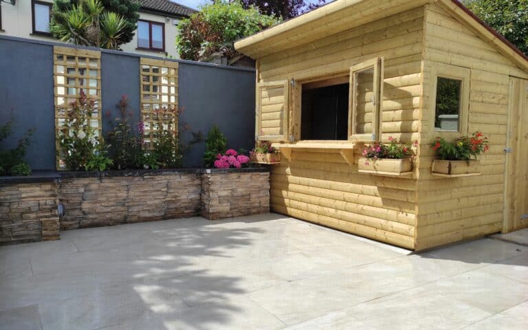 A tidy patio with light stone tiles, a raised garden bed with flowers and shrubs along a grey wall, and a wooden shed with open windows and flower boxes, under a partly sunny sky. A house and trees are visible in the background.