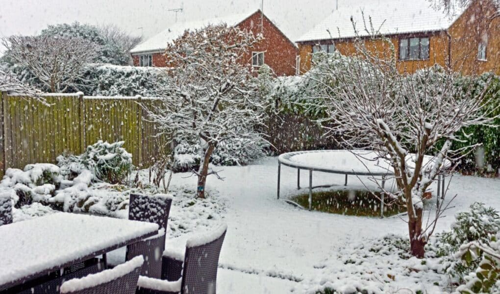 A back garden covered in fresh snow shows garden furniture, bare trees, bushes, a round trampoline, and two brick houses in the background. Snow is actively falling, blanketing the scene in white and creating a peaceful winter atmosphere.