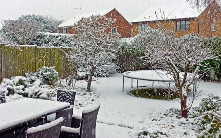 A back garden covered in fresh snow shows garden furniture, bare trees, bushes, a round trampoline, and two brick houses in the background. Snow is actively falling, blanketing the scene in white and creating a peaceful winter atmosphere.