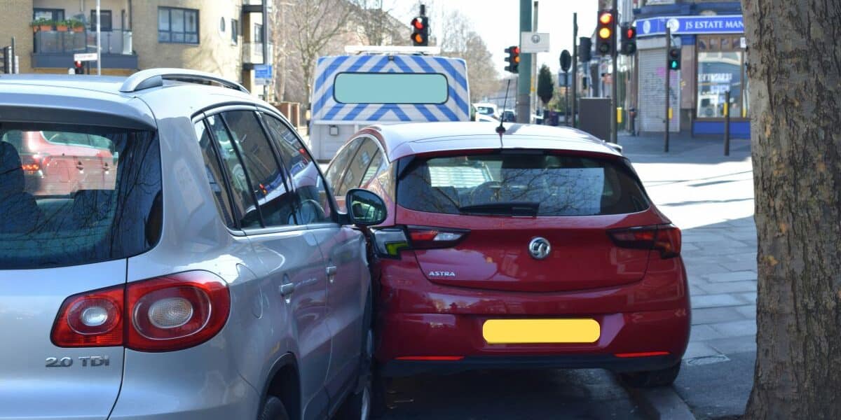 A silver SUV and a red hatchback are parked closely on a city street, their bumpers nearly touching. A police van is parked ahead, and traffic lights, buildings, and a tree line the pavement on a sunny day. Number plates are blurred for privacy.