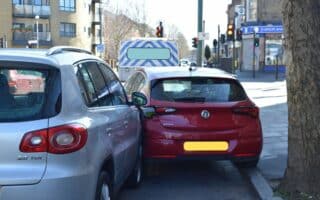 A silver SUV and a red hatchback are parked closely on a city street, their bumpers nearly touching. A police van is parked ahead, and traffic lights, buildings, and a tree line the pavement on a sunny day. Number plates are blurred for privacy.
