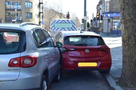 A silver SUV and a red hatchback are parked closely on a city street, their bumpers nearly touching. A police van is parked ahead, and traffic lights, buildings, and a tree line the pavement on a sunny day. Number plates are blurred for privacy.