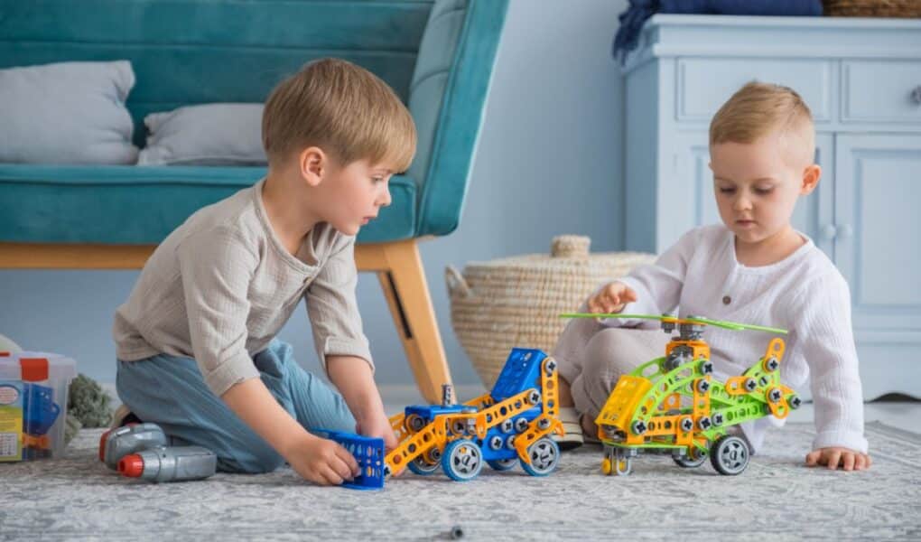Two young children sit on a rug indoors, playing with colourful construction toys—a blue car and a yellow-green helicopter. The children focus intently on their toys. A blue sofa, woven basket, and blue furniture appear in the background.