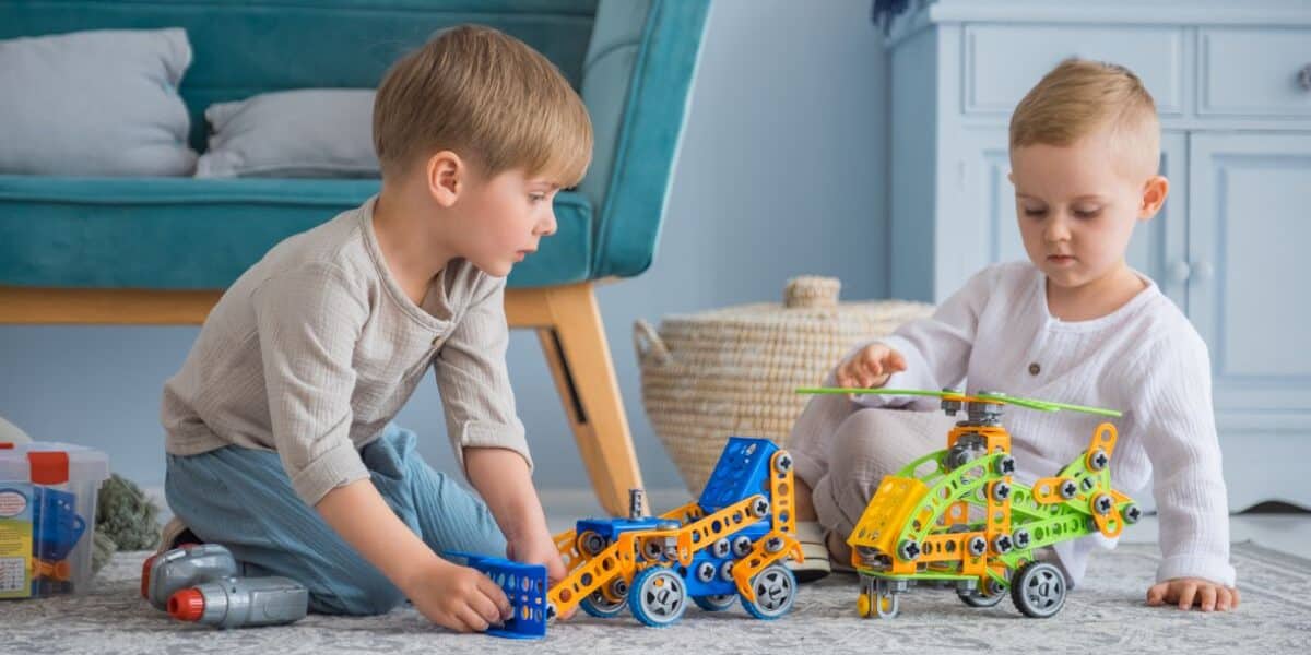 Two young children sit on a rug indoors, playing with colourful construction toys—a blue car and a yellow-green helicopter. The children focus intently on their toys. A blue sofa, woven basket, and blue furniture appear in the background.