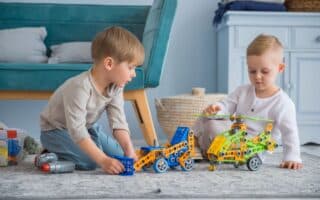 Two young children sit on a rug indoors, playing with colourful construction toys—a blue car and a yellow-green helicopter. The children focus intently on their toys. A blue sofa, woven basket, and blue furniture appear in the background.