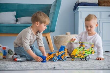 Two young children sit on a rug indoors, playing with colourful construction toys—a blue car and a yellow-green helicopter. The children focus intently on their toys. A blue sofa, woven basket, and blue furniture appear in the background.