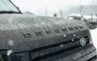 Close-up of a dark Land Rover Defender's wet bonnet, with large raindrops covering the surface. The word "DEFENDER" is visible in raised letters, and the Land Rover logo appears on the right side. Snowflakes are falling and the background is blurred.