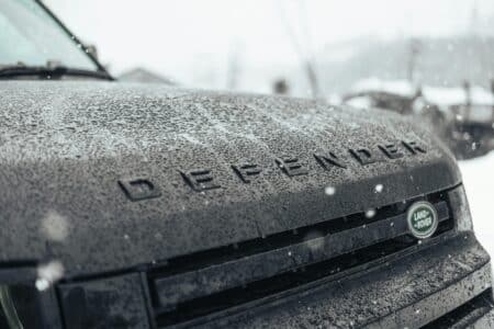 Close-up of a dark Land Rover Defender's wet bonnet, with large raindrops covering the surface. The word "DEFENDER" is visible in raised letters, and the Land Rover logo appears on the right side. Snowflakes are falling and the background is blurred.