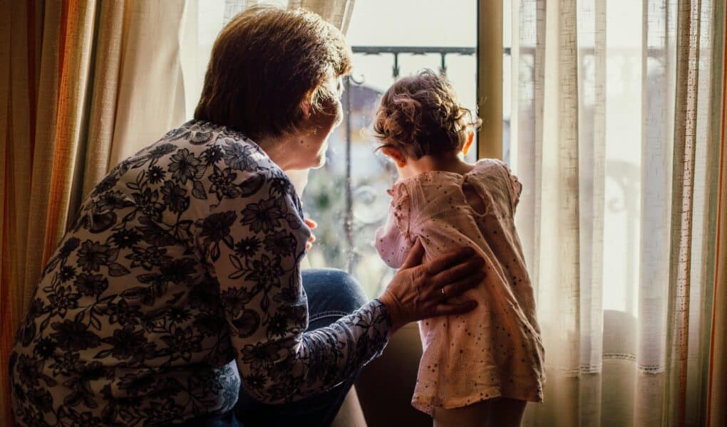 An older woman with short hair and a floral shirt gently holds a young child in a pink dress, both looking out of a bright window framed by sheer curtains. Sunlight softly illuminates them from behind, creating a warm, intimate atmosphere.