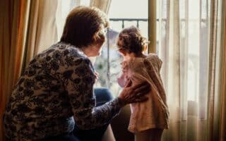 An older woman with short hair and a floral shirt gently holds a young child in a pink dress, both looking out of a bright window framed by sheer curtains. Sunlight softly illuminates them from behind, creating a warm, intimate atmosphere.