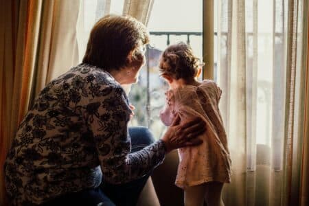 An older woman with short hair and a floral shirt gently holds a young child in a pink dress, both looking out of a bright window framed by sheer curtains. Sunlight softly illuminates them from behind, creating a warm, intimate atmosphere.