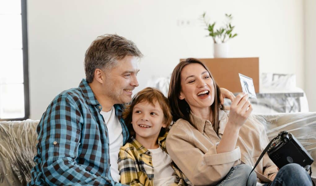 A smiling family of three sits on a sofa. The man and woman, both casually dressed, sit on either side of a young boy. The woman holds up a photo while the others look at it, and a camera rests on her lap. A potted plant and boxes are in the background.