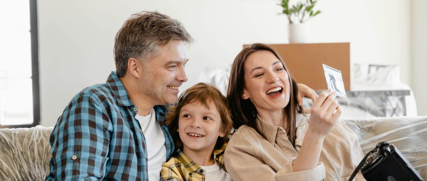 A smiling family of three sits on a sofa. The man and woman, both casually dressed, sit on either side of a young boy. The woman holds up a photo while the others look at it, and a camera rests on her lap. A potted plant and boxes are in the background.