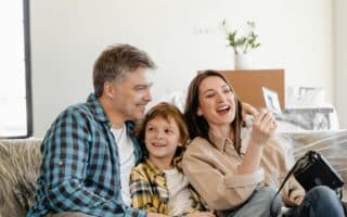 A smiling family of three sits on a sofa. The man and woman, both casually dressed, sit on either side of a young boy. The woman holds up a photo while the others look at it, and a camera rests on her lap. A potted plant and boxes are in the background.