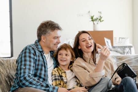 A smiling family of three sits on a sofa. The man and woman, both casually dressed, sit on either side of a young boy. The woman holds up a photo while the others look at it, and a camera rests on her lap. A potted plant and boxes are in the background.