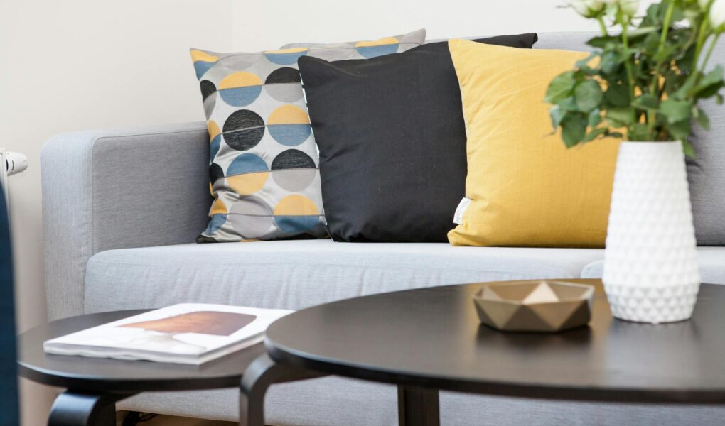 A modern living room features a light grey sofa with three cushions—one patterned with geometric shapes in yellow, grey, and black; one solid black; and one solid yellow. In front, two black tables hold a white vase of white roses, a small golden bowl, and a magazine.